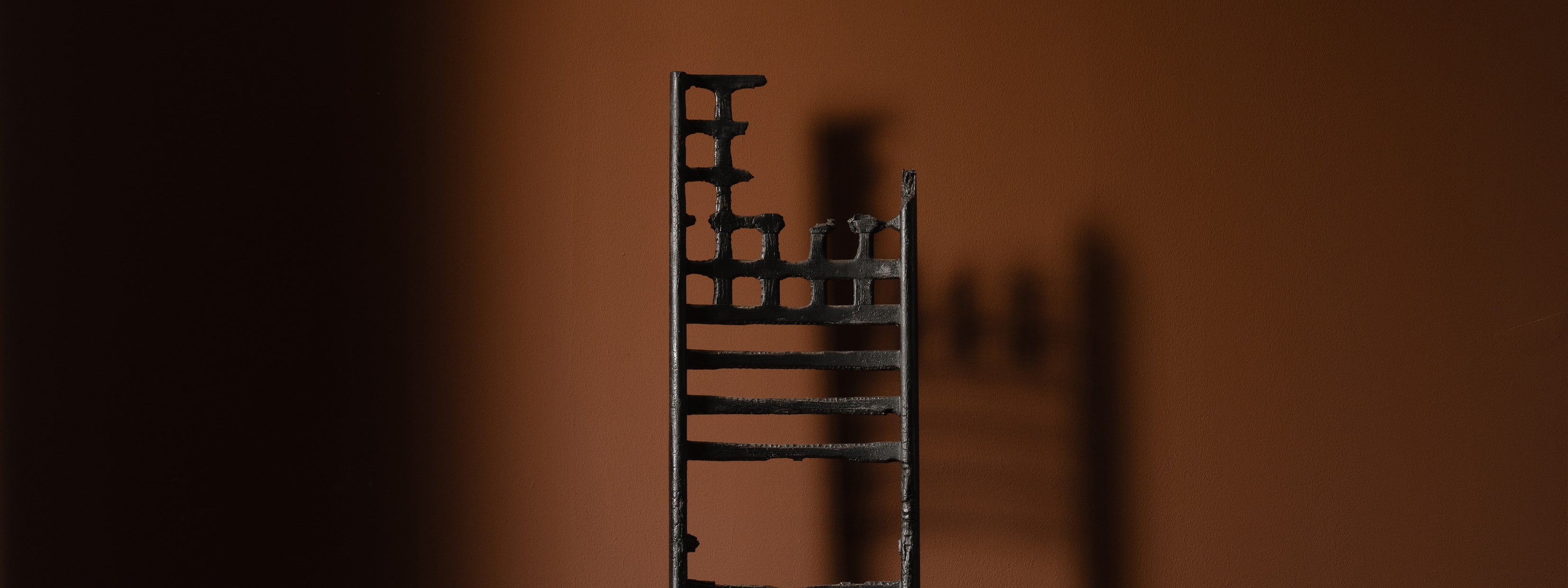 A close-up of a tall, narrow black metal sculpture resembling a ladder with uneven, abstract rungs. The piece casts a shadow on a warm brown wall, creating depth and contrast in the minimalist composition.