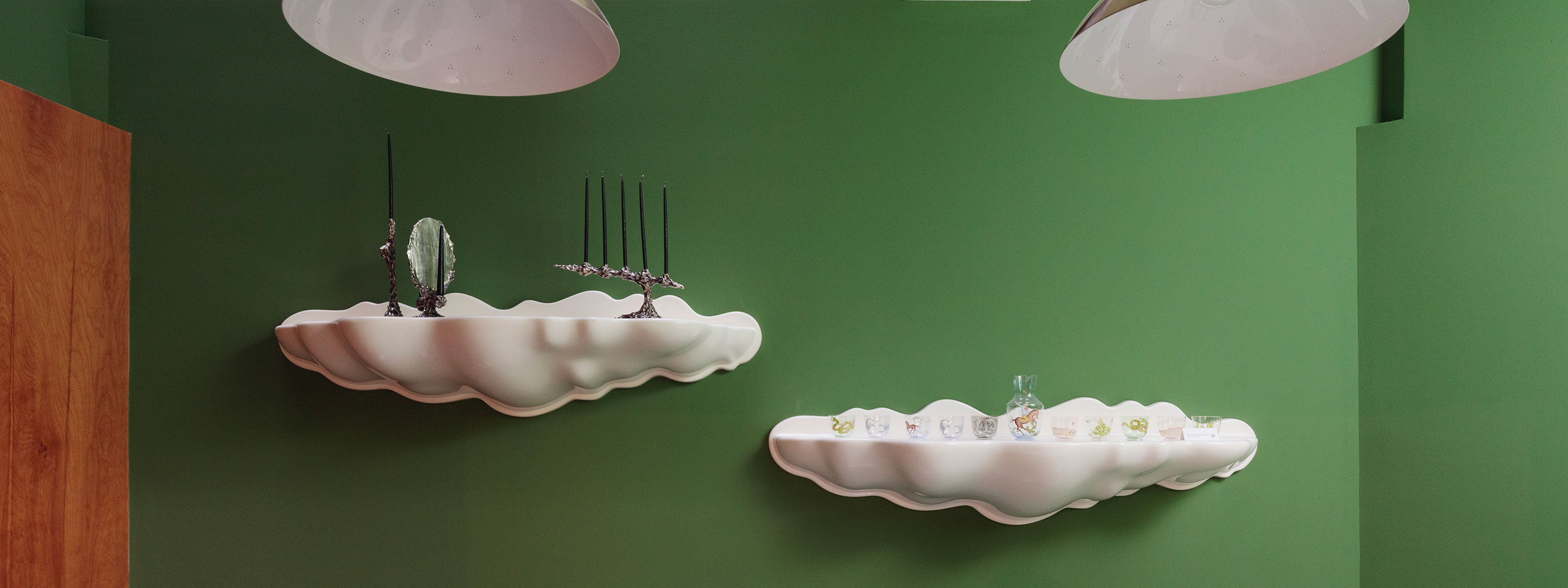 A modern green-walled interior featuring two sculptural white shelves shaped like clouds. The upper shelf displays black candlesticks and decorative objects, while the lower shelf holds colorful glassware and a clear vase. Large overhead pendant lights partially appear at the top of the image, adding to the contemporary design.