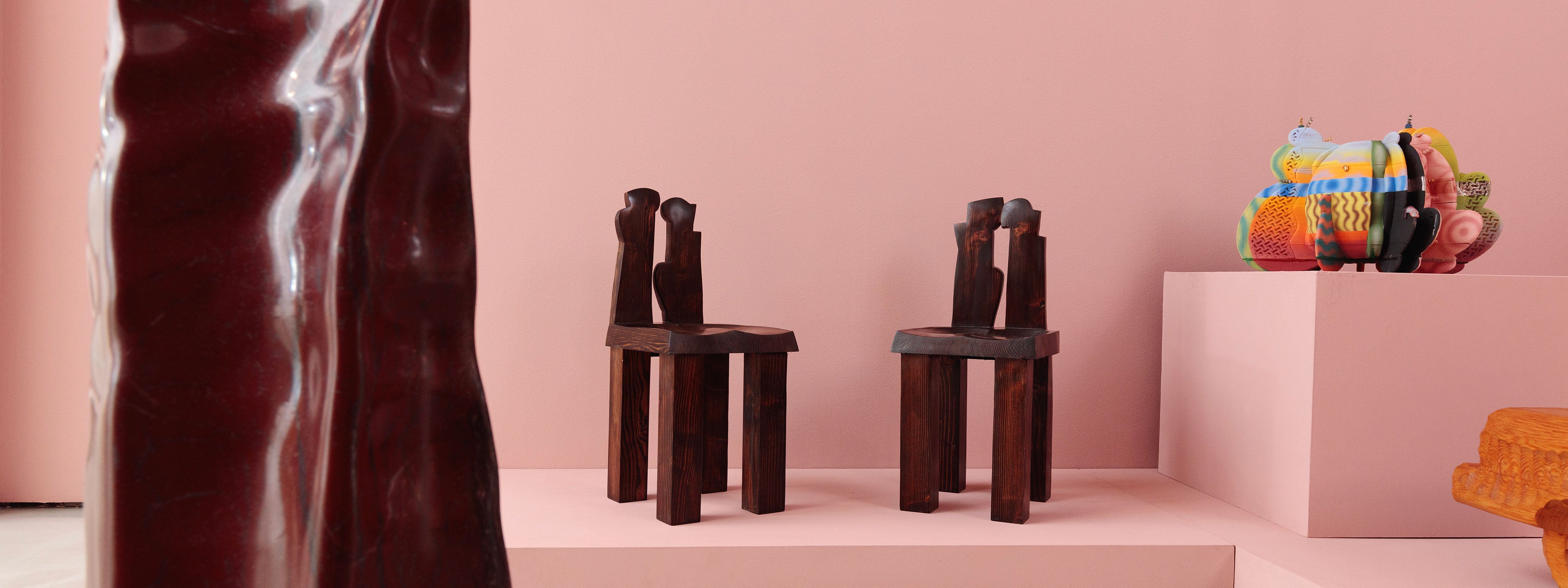 A modern interior with Alkemis Paint blush-pink walls featuring two sculptural dark wood chairs with abstract backrests on a raised platform. In the background, a colorful, rounded abstract sculpture rests on a pedestal, while a glossy burgundy form partially obscures the left side of the image. The overall aesthetic is artistic and contemporary.