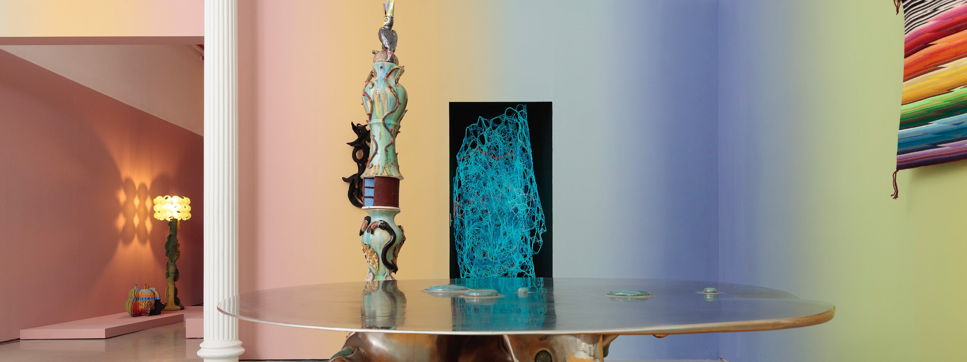 A contemporary gallery space with gradient walls shifting from pink to blue to yellow. At the center, a tall sculptural column with ornate ceramic elements rises through a reflective round table. In the background, a glowing sculptural lamp with a green base and flower-like yellow shade is placed against a pink wall, while a colorful woven textile hangs on the right.