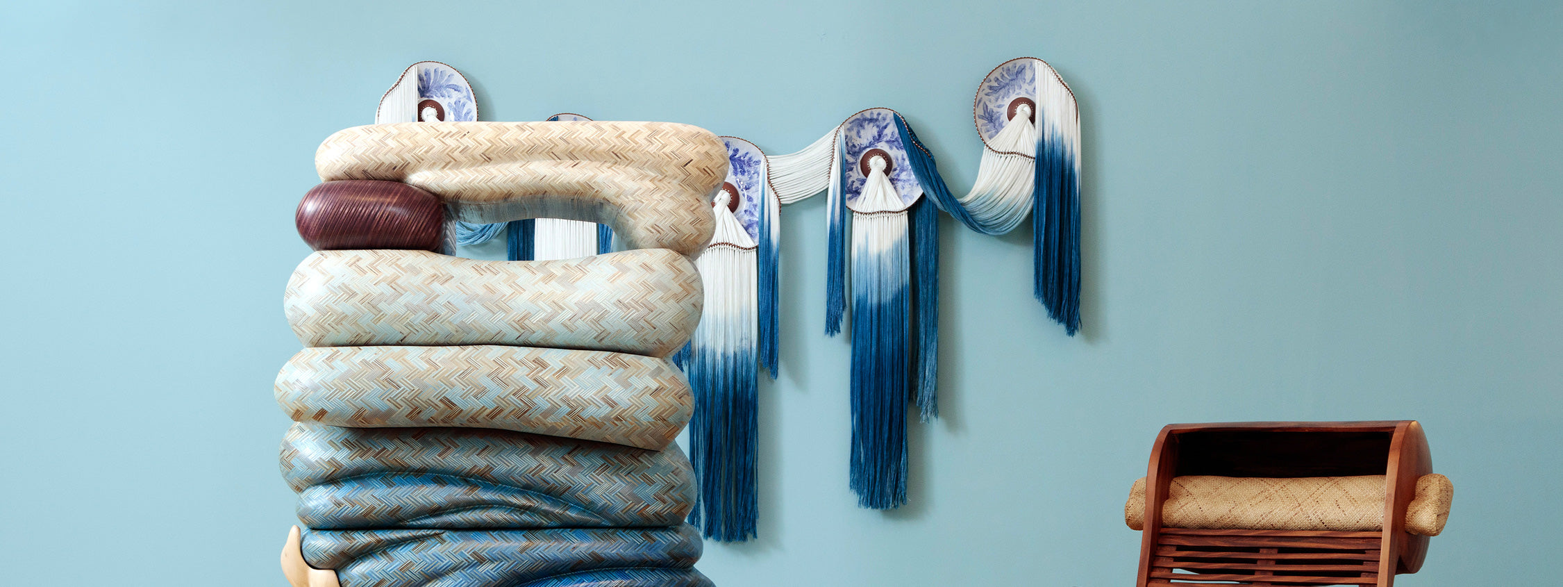 A sculptural piece made of stacked, coiled cushion-like forms in woven beige and blue patterns stands against a pale blue wall. Behind it, decorative ceramic plates with blue designs and long ombre tassels hang in a draped formation. To the right, a wooden chair with woven seating is partially visible.